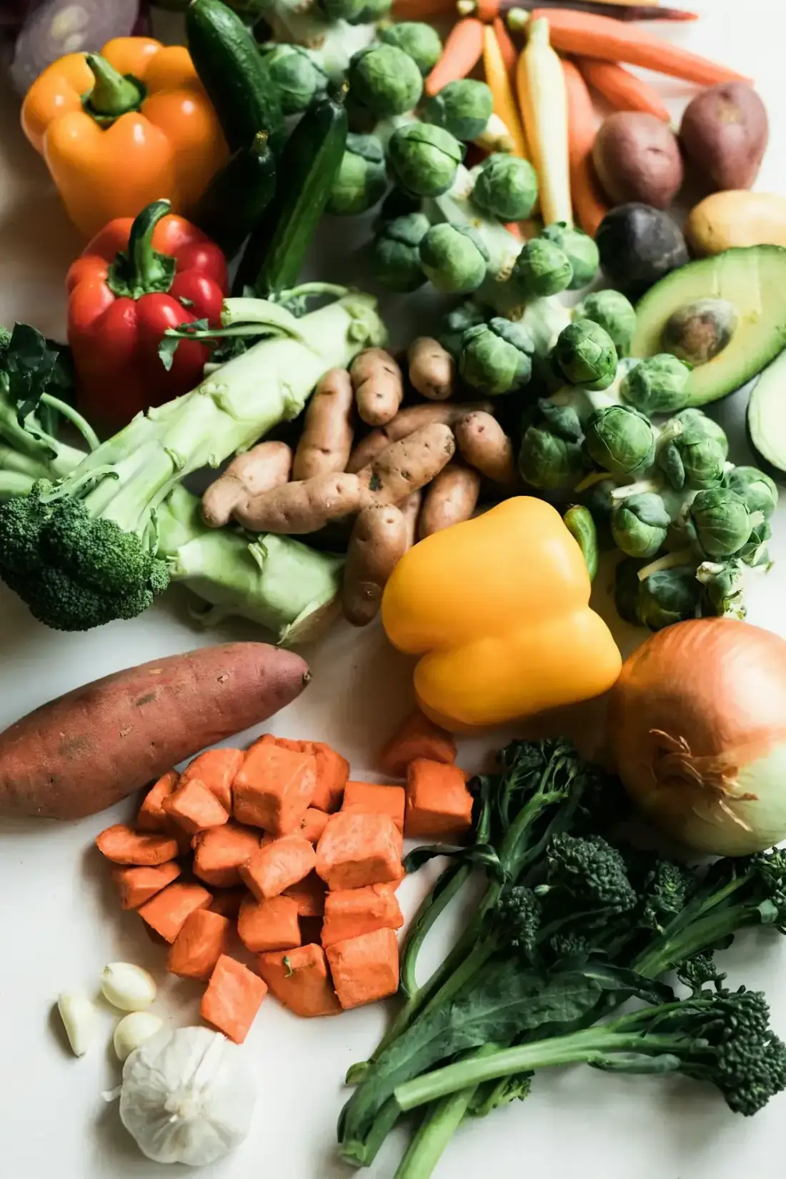 Legumes frescos sobre superfície branca: brócolis, batata-doce, pimentão amarelo e cebola