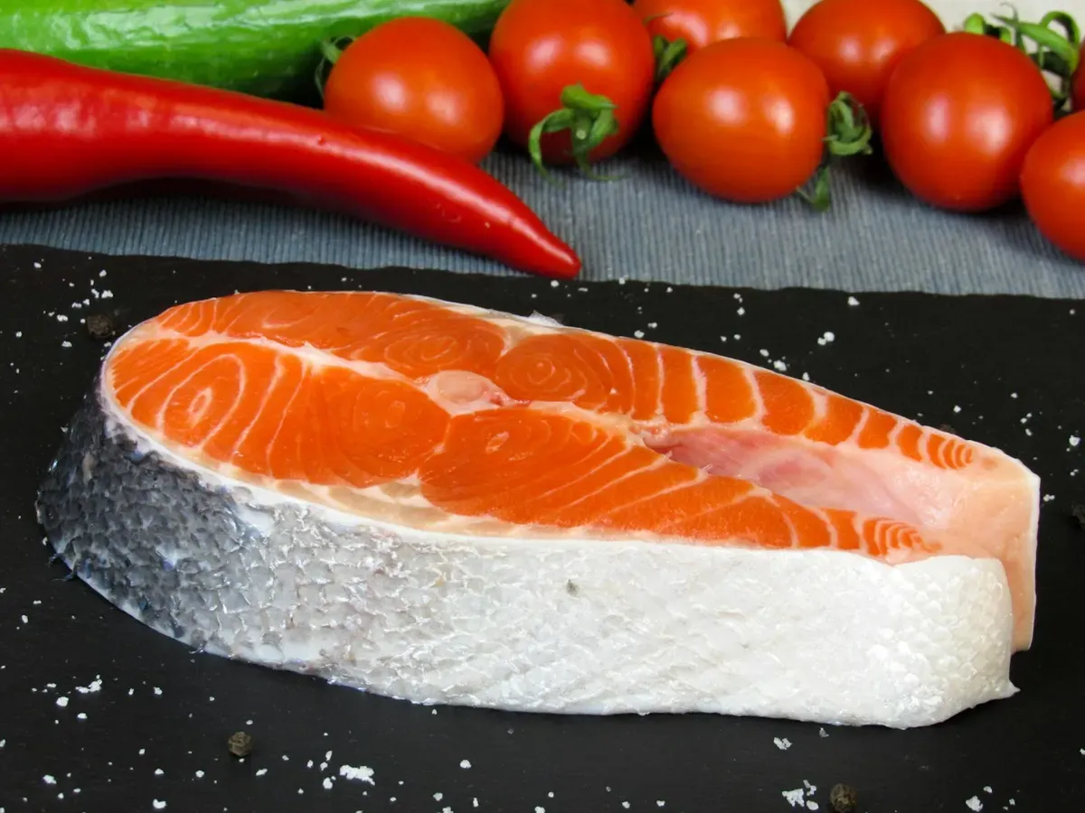 Posta de salmão cru sobre pedra escura com tomates e pimenta ao fundo